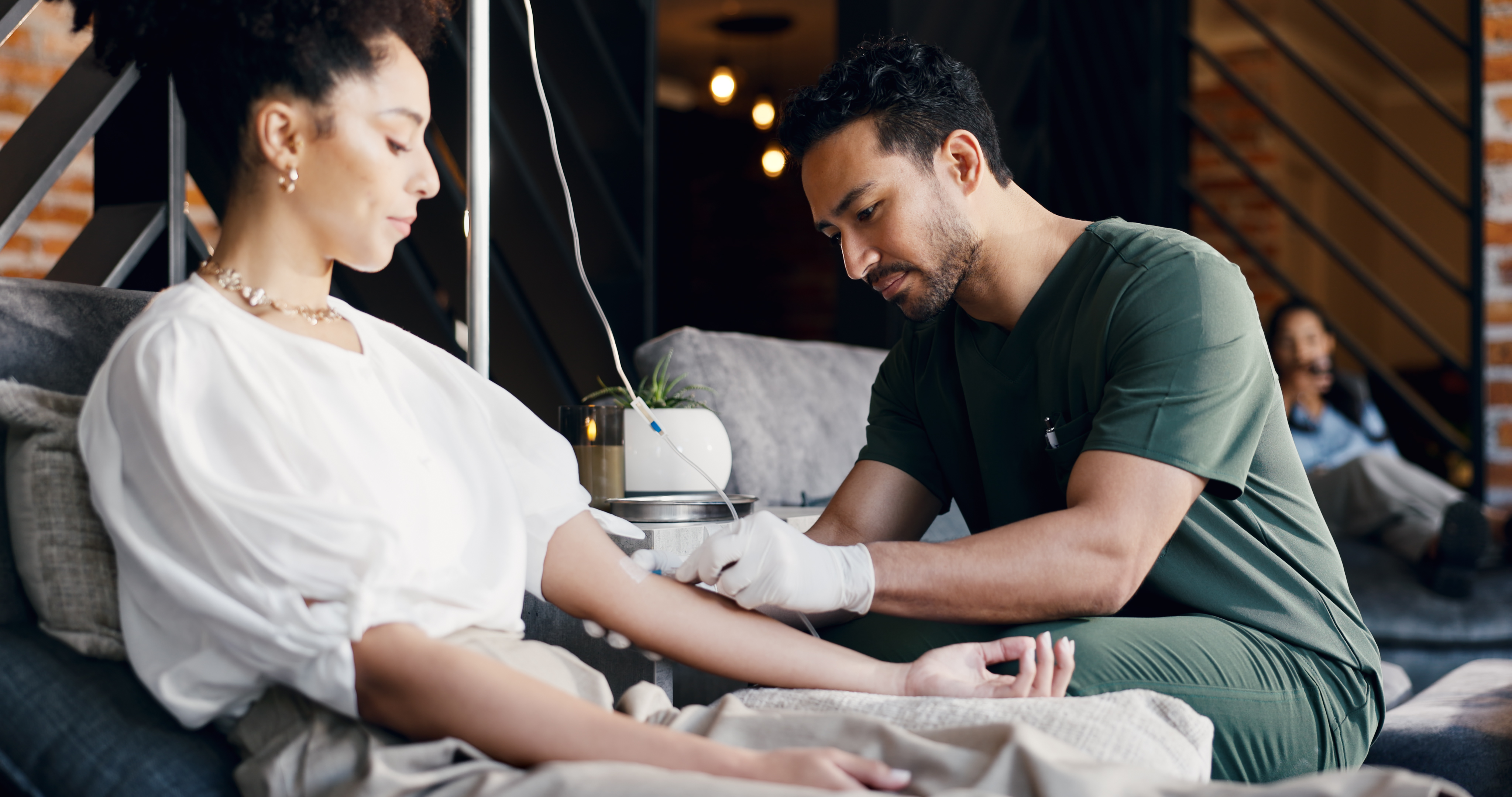 A nurse in green scrubs administering an IV drip to a relaxed woman in a calm spa-like studio