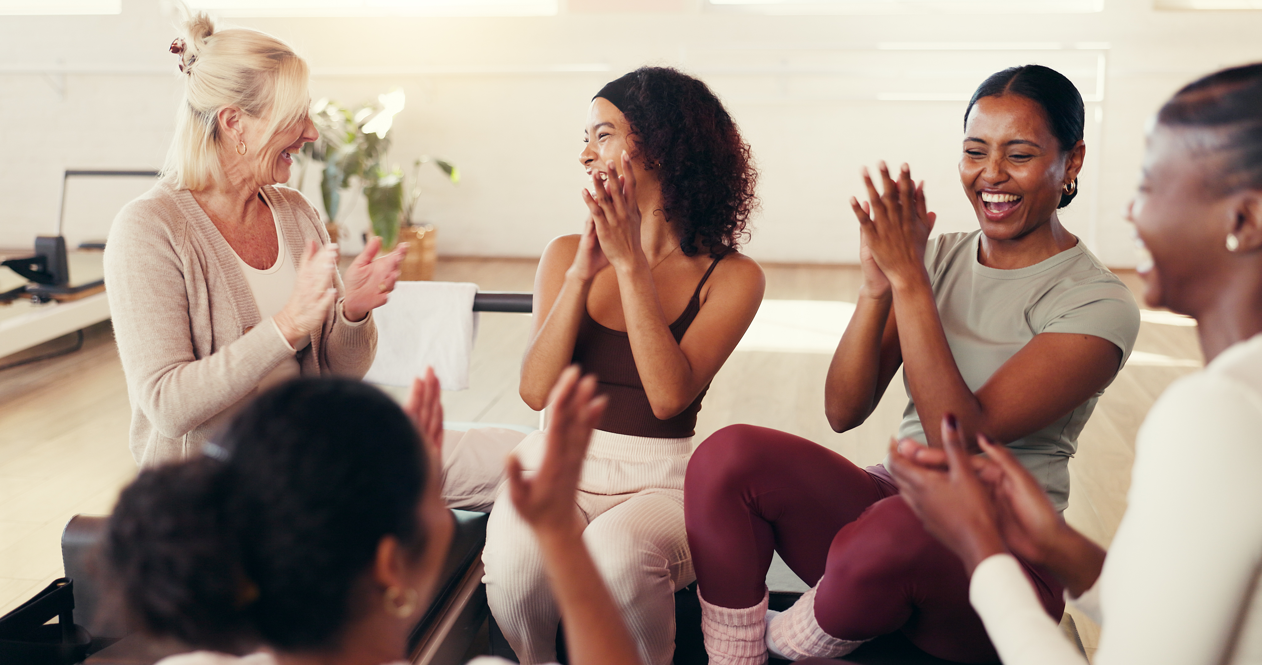 A diverse group of women laughing and clapping together in a bright wellness studio